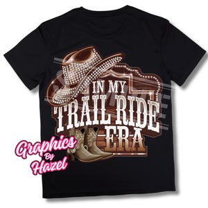 Peut inclure: T-shirt noir avec un chapeau de cowboy marron orné de strass, des bottes de cowboy marron et une pancarte en bois sur laquelle est inscrit "In My Trail Ride Era".