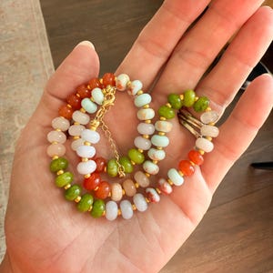 May include: A multi-strand necklace featuring a variety of colorful beads in shades of green, orange, pink, and white. The beads are strung together with gold-colored spacers and a delicate gold chain.