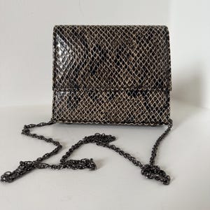 May include: A small, rectangular crossbody bag with a snakeskin pattern in shades of brown and beige. It has a dark metal chain strap. The bag is set against a white background.