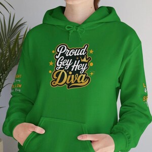 Op de afbeelding: Een groene hoodie met de tekst "Proud Gey Hey Diva" in zwart en goud. De sweatshirt heeft een voorzak en een capuchon met trekkoord. Extra tekst is zichtbaar op de linkermouw.