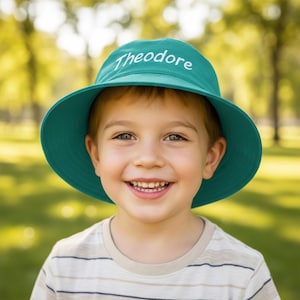 May include: A teal bucket hat featuring the name Theodore embroidered in white script lettering across the front crown. The hat has a wide, downward-sloping brim and a structured dome top, providing a solid colour design with clear text.