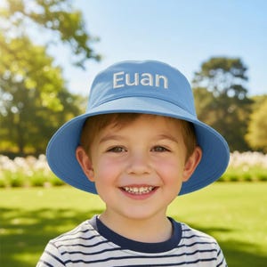 May include: A light blue bucket hat featuring the name Euan embroidered in white lettering on the front. The hat has a wide, downward-sloping brim and a structured crown, providing a simple and functional design for sun protection.
