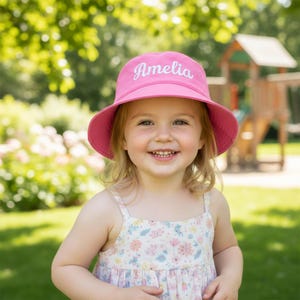 May include: A bright pink bucket hat featuring the name Amelia embroidered in white script lettering across the front. The hat has a wide, downward-sloping brim and a structured crown, providing a simple and functional design for sun protection.
