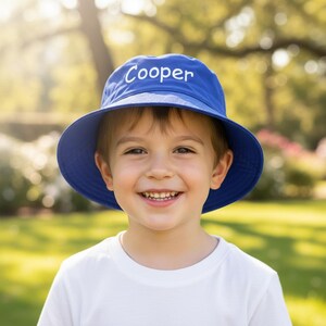 May include: A solid royal blue bucket hat featuring the name Cooper embroidered in white thread across the front crown. The hat has a wide, downward-sloping brim and a rounded top, providing a simple and functional design for sun protection.