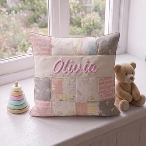 May include: A square patchwork cushion featuring a central cream-coloured horizontal band embroidered with the name Olivia in pink script. The cushion is decorated with various pastel-coloured fabric squares displaying patterns of giraffes, elephants, and lions.