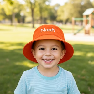 May include: A bright orange bucket hat featuring the name Noah embroidered in white thread across the front crown. The hat has a wide, downward-sloping brim and a rounded top, providing a simple and functional design for sun protection.