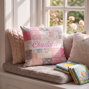 May include: A square patchwork cushion featuring various shades of pink, white, and grey fabric squares with patterns of animals and chevrons. The name Charlotte is embroidered in pink script across the centre of the cushion, which is placed among other cushions.