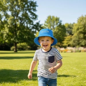 May include: A bright blue bucket hat with the name Archie embroidered in white thread on the front, paired with a short-sleeved white t-shirt featuring thin horizontal navy blue stripes and a small chest pocket, worn with blue denim jeans.