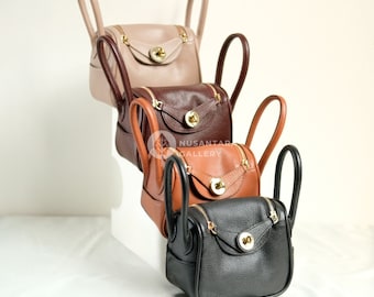 May include: Four leather handbags in different colors, stacked on top of each other. The top bag is beige, the second is brown, the third is tan, and the bottom bag is black.