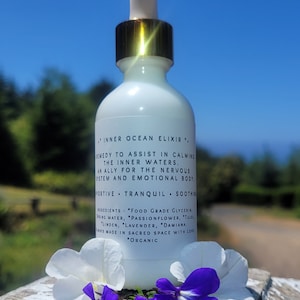 以下が含まれることがあります： 「インナーオーシャンエリクサー」とラベルが貼られた、金色の蓋とスポイトが付いた白いボトル。ボトルは、紫と白の花が咲いた木製の表面の上に置かれています。ラベルには、「内なる水を落ち着かせるのを助けるための薬。神経系と感情的な体の味方。サポート、穏やか、鎮静。成分：食品グレードのグリセリン、湧き水、パッションフラワー、トゥルシー、リンデン、ラベンダー、ダミアナ。常に神聖な空間で愛を込めて作られています。*オーガニック」と書いてあります。