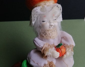 Puede incluir: Una pequeña muñeca de porcelana con un vestido de encaje blanco y un sombrero de fieltro naranja, sosteniendo una pequeña calabaza naranja. La muñeca está sentada sobre una calabaza naranja más grande con un tallo verde.