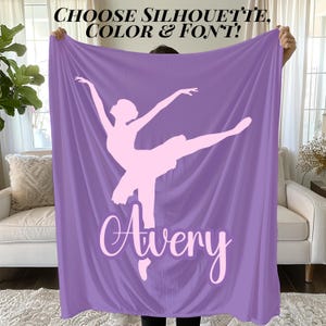 May include: A lavender blanket featuring a light pink ballerina silhouette and the name "Avery" in a cursive font. The blanket has the text "CHOOSE SILHOUETTE, COLOR & FONT!" at the top. The blanket is held up in front of a window and a white couch.