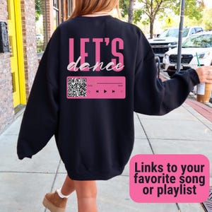 Puede incluir: Sudadera negra con un gráfico rosa "Let's Dance" y un código QR. El texto "Enlaces a tu canción o lista de reproducción favorita" es visible debajo del gráfico.