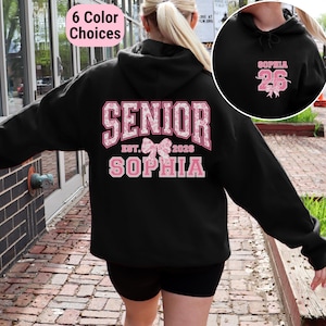 May include: Black hooded sweatshirt with pink floral "SENIOR" design, "EST. 2026" and "SOPHIA" text. A smaller image shows "SOPHIA 26" on the hood. The image also shows "6 Color Choices" in a pink box.
