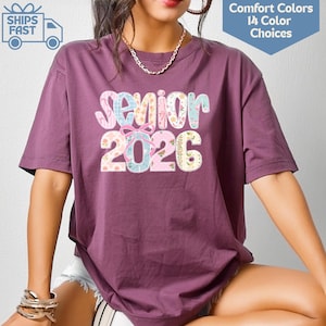 以下が含まれることがあります： 鮮やかな模様のフォントで「Senior 2026」と書かれた、プラム色の半袖Tシャツ。シャツには「Comfort Colors 14 Color Choices」と書かれたグラフィックも表示されています。