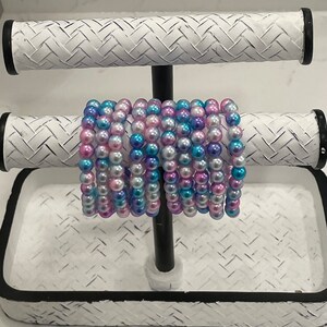 May include: A stack of pearl bracelets in shades of pink, blue, and white on a white display stand.