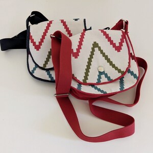 Könnte beinhalten: Zwei Stoff-Umhängetaschen mit einem geometrischen Muster in Rot-, Grün- und Blautönen. Die Tasche links hat einen schwarzen Riemen, die Tasche rechts einen roten Riemen.