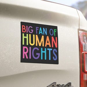 May include: A black rectangular bumper sticker with the text "BIG FAN OF HUMAN RIGHTS" in rainbow-colored letters is affixed to the back of a white vehicle. The vehicle has a 4x4 emblem.