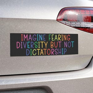 May include: A rectangular black bumper sticker with the text "IMAGINE FEARING DIVERSITY BUT NOT DICTATORSHIP" in rainbow-colored letters. The sticker is affixed to the rear of a silver car.