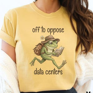 May include: A mustard yellow short-sleeve t-shirt featuring a graphic of a green frog wearing a brown hat and carrying a backpack while reading a book. The text above the frog reads off to oppose and the text below the frog reads data centers.