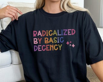 May include: A black short-sleeved t-shirt featuring the multi-colored text "RADICALIZED BY BASIC DECENCY" printed on the front in a playful, rounded font, accompanied by small four-pointed star icons to the right of the lettering.