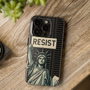 May include: A black phone case featuring an illustration of the Statue of Liberty holding a sign that reads "RESIST." The background has a repeating pattern of the words "fuck trump." The case is on a wooden surface.