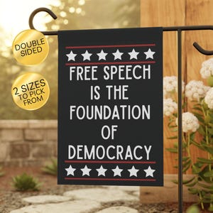May include: A rectangular black garden flag featuring the white text "FREE SPEECH IS THE FOUNDATION OF DEMOCRACY" centered between two horizontal red lines and rows of white stars at the top and bottom, hanging on a black metal stand.