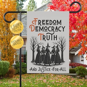 May include: A double-sided garden flag with the words "Freedom, Democracy, Truth, And Justice For All" in black and orange. The flag features a graphic of six women in witch hats and dresses holding hands. The flag is displayed on a black metal stand.