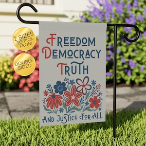May include: A double-sided garden flag with patriotic text and floral design. The flag displays the words 'Freedom, Democracy, Truth, and Justice For All' in red and blue. A floral pattern of red and blue flowers is at the bottom. The flag is hanging on a black metal stand.
