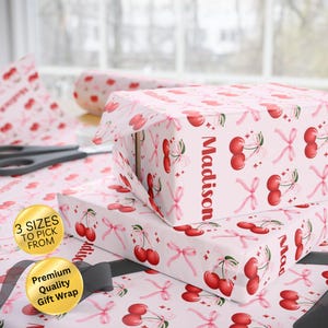 May include: Gift wrap featuring a repeating pattern of red cherries and pink bows on a light pink background. The word "Madison" is printed vertically on the side of the wrapped gift. The image also includes scissors and a roll of wrapping paper.