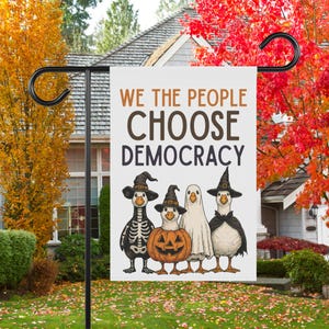 May include: A white garden flag with the text "WE THE PEOPLE CHOOSE DEMOCRACY" in brown. The flag features four cartoon geese dressed in Halloween costumes: a skeleton, a pumpkin, a ghost, and a witch.