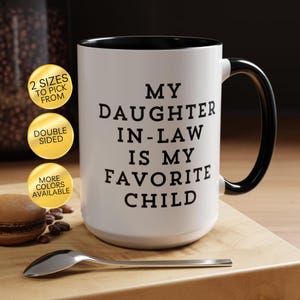 May include: A white ceramic mug with a black interior and a black handle. The mug features black text that reads: MY DAUGHTER IN-LAW IS MY FAVORITE CHILD. Three gold circular badges are present, stating 2 SIZES TO PICK FROM, DOUBLE SIDED, and MORE COLORS AVAILABLE.