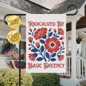 May include: A double-sided garden flag with the text "Radicalized by Basic Decency" in red script. The flag features a floral design with red flowers and blue leaves. The flag is displayed on a black metal stand.