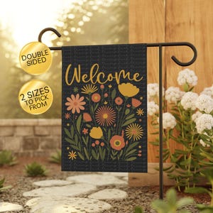 May include: A double-sided garden flag with a black background and the word "Welcome" in gold script. The flag features a floral design in shades of orange, peach, and yellow. The flag is displayed on a black metal stand in a garden setting.