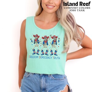 May include: A light mint green tank top featuring a graphic of three skeletons wearing red, white, and blue cowboy hats, vests, and boots. The skeletons are surrounded by colorful stars, with the text FREEDOM DEMOCRACY TRUTH printed below.