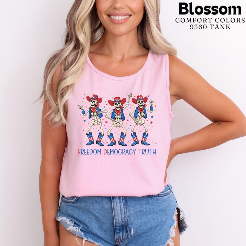 May include: A light pink tank top featuring a graphic of three skeletons wearing red, white, and blue cowboy hats, vests, and boots. The text "FREEDOM DEMOCRACY TRUTH" is printed below the skeletons, accompanied by small red and blue stars.
