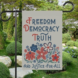 May include: A garden flag with the words "Freedom, Democracy, Truth, And Justice For All" in red and blue lettering. The flag features a floral design with red and blue flowers. The flag is hanging on a black metal stand in a garden setting.