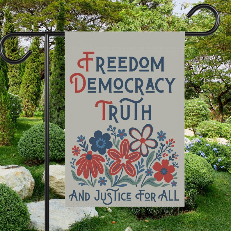 May include: A garden flag with the words "Freedom, Democracy, Truth, And Justice For All" in red and blue lettering. The flag features a floral design with red and blue flowers. The flag is hanging on a black metal stand in a garden setting.