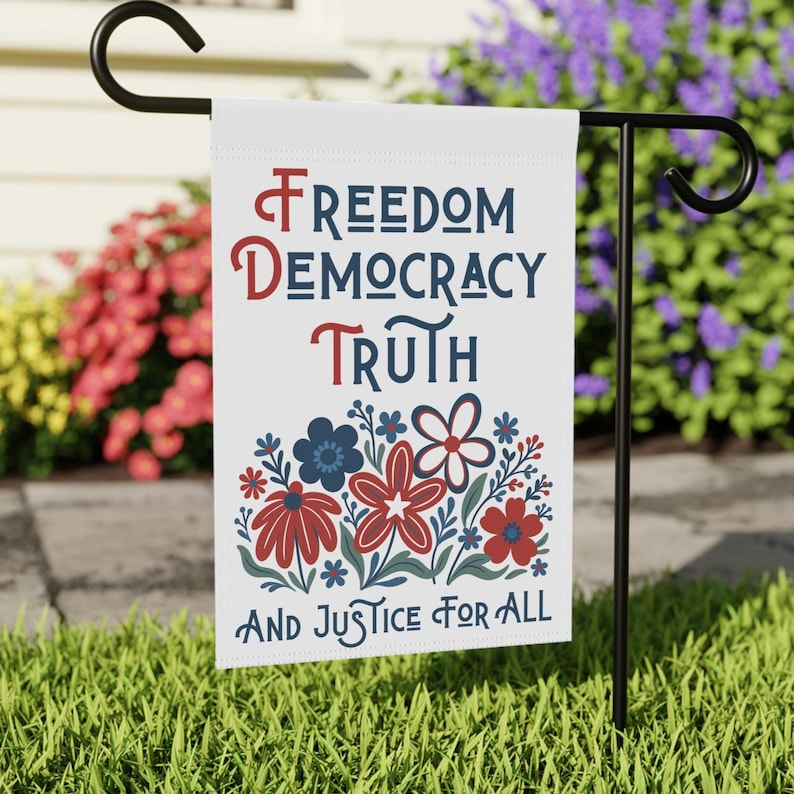 May include: A white garden flag with the words 'Freedom, Democracy, Truth, And Justice For All' in navy and red lettering. Below the words is a floral design with red, white, and navy flowers. The flag is displayed on a black metal garden stake.