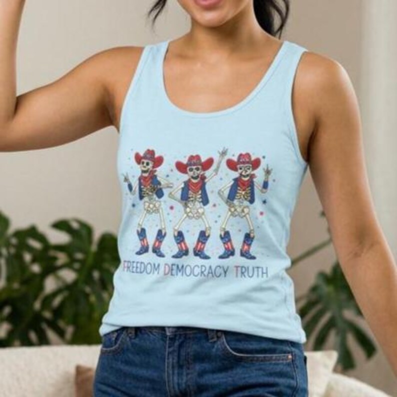 May include: A light blue tank top featuring a graphic of three skeletons wearing red cowboy hats, blue vests, and red, white, and blue cowboy boots. The skeletons are dancing, and the text below them reads FREEDOM DEMOCRACY TRUTH in colorful letters.