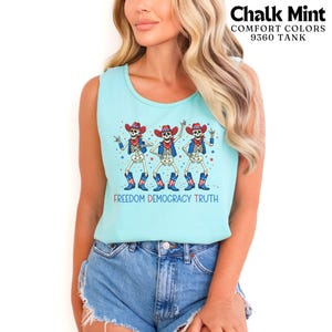 May include: A light mint green tank top featuring a graphic of three skeletons wearing red, white, and blue cowboy hats, vests, and boots. The text "FREEDOM DEMOCRACY TRUTH" is printed below the skeletons, with small stars scattered around them.