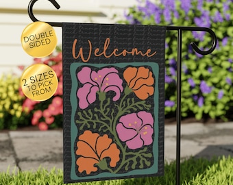 May include: A double-sided garden flag with a floral design and the word "Welcome" in orange script. The flowers are pink and orange, set against a dark background with a green border. The flag is displayed on a black metal stand.