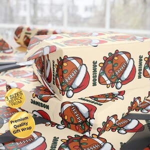 May include: Gift wrap featuring a repeating pattern of a football wearing a Santa hat and string lights. The name "JAMISON" is printed on the wrap. The wrap is cream-colored and has the text "3 SIZES TO PICK FROM" and "Premium Quality Gift Wrap".