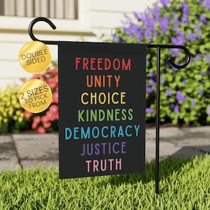 May include: A black garden flag with the words "FREEDOM, UNITY, CHOICE, KINDNESS, DEMOCRACY, JUSTICE, TRUTH" in rainbow colors. The flag is double-sided and available in two sizes. It is displayed on a black metal stand in a garden.