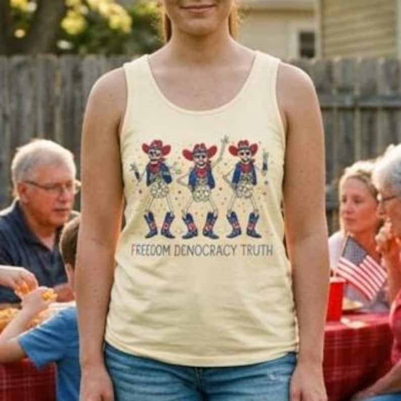 May include: A light yellow tank top featuring a graphic of three skeletons wearing cowboy hats, vests, and boots, with red and blue accents. Below the skeletons, the text reads FREEDOM DENOCRACY TRUTH in a bold, dark font, centered on the chest.