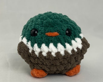 May include: A green and brown crocheted plush toy shaped like a bird with white accents. The toy has black eyes and an orange beak.