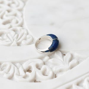 Könnte beinhalten: Ein silberner Ring mit einem breiten Band und einem dunkelblauen Steinbesatz, möglicherweise Lapislazuli. Der Ring liegt auf einer weißen, geschnitzten Oberfläche, die den Kontrast hervorhebt.