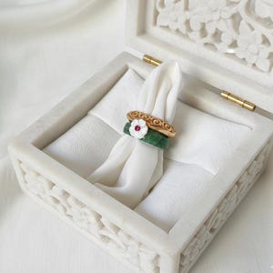 May include: A white marble jewelry box with carved floral designs, holding a white cloth with three rings. The rings include a gold band with a floral design, a green band, and a green band with a white flower.