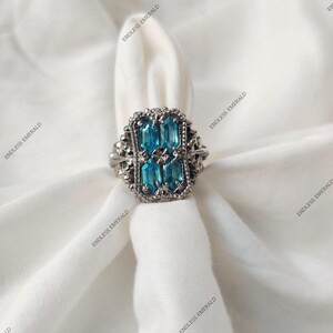 Könnte beinhalten: Ein silberfarbener Ring mit rechteckiger Fassung, besetzt mit vier rechteckigen, hellblauen Edelsteinen und einem zentralen Edelstein. Der Ring hat eine verzierte, detaillierte Fassung und wird auf weißem Stoff präsentiert.
