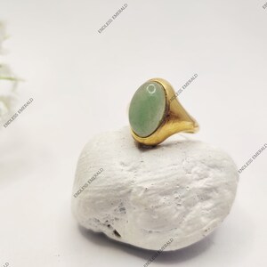 Könnte beinhalten: Ein goldener Ring mit einem hellgrünen ovalen Edelstein. Der Ring liegt auf einem weißen Stein.